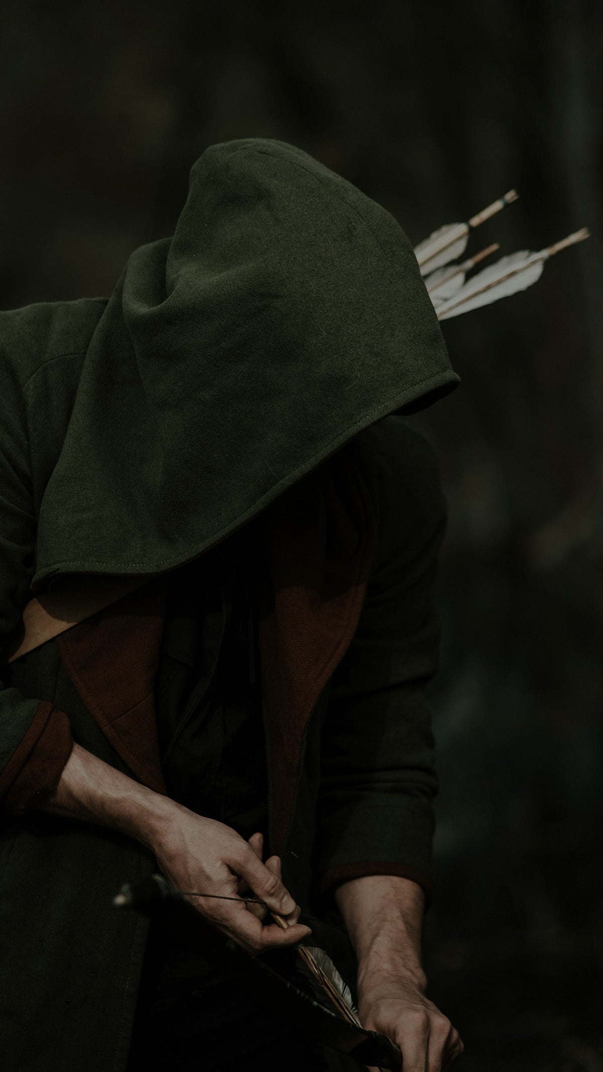 Hooded figure standing in a forest with a bow, evoking the stealth and resilience of the Ranger collection by Fabled Fox Clothiers.